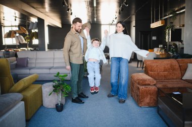 Ebeveynler, mobilya galerisini keşfederken mutlu bir çocuğu elleriyle kaldırıyorlar, evleri için yeni eşyalar seçiyorlar.