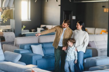 Genç bir aile, anne, baba ve oğul, mobilya galerisinde birlikte vakit geçiriyorlar, yeni bir kanepe arıyorlar.