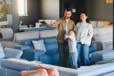 Mobilya galerisindeki anne ve babalar rahatlığı test ederken gülümseyerek ve parmakla göstererek ev içleri için yeni bir kanepe seçiyorlar.
