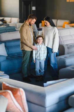Ebeveynler ve küçük oğulları gülümserken parlak bir sergi salonunda yeni bir kanepe seçiyorlar. Aile alışverişi ve ev planlamasının keyfini çıkarıyorlar.