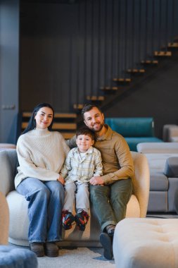 Üç kişilik mutlu bir aile, modern bir galeride yeni mobilya alırken bir koltukta oturup gülümsüyor.