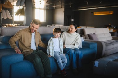 Modern bir mobilya galerisinde mavi bir koltukta oturan mutlu üç kişilik aile, yeni ev mobilyalarını seçiyorlar.