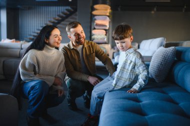 Ebeveynler ve oğulları modern bir sergi salonunda rahat bir kanepe seçiyorlar, ev geliştirme ve aile yaşam tarzına odaklanıyorlar.