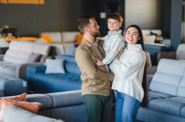 Genç bir aile, gülümseyen ebeveynler ve küçük bir çocuk, geniş bir mobilya mağazasında rahat modern koltukları seçiyorlar.