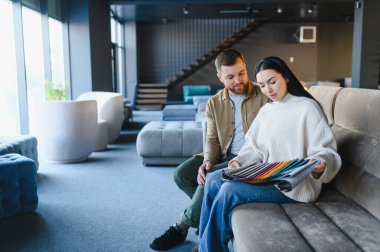 Genç bir çift kanepede oturmuş çeşitli kumaş örneklerine bakıyor ve ev dekorasyonu seçeneklerini tartışıyorlar.