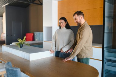 Genç çift mutfak tasarım seçeneklerini ve malzemelerini araştırıyor, çağdaş bir mobilya mağazasında ahşap bir masa ve tezgaha dokunuyor.