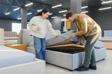 Modern bir sergi salonunda açık depolama bölmesi olan bir yatağı inceleyen çift, ev eşyaları ve seçime odaklanıyor.