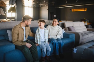 Mutlu bir aile mobilya mağazasını keşfediyor, mavi bir koltukta oturuyor ve yeni mobilyaları seçerken birlikte gülüyor.