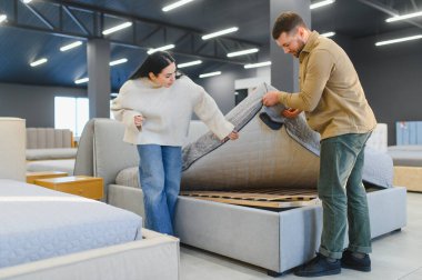 Genç çift yeni bir yatak almak için alışveriş yapıyor, modern bir mobilya galerisinde çerçeveyi incelemek için yatağı kaldırıyor.