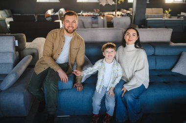 Modern bir mobilya dükkanında mavi bir koltukta oturmuş gülümseyen genç bir aile evleri için yeni parçalar seçiyor.