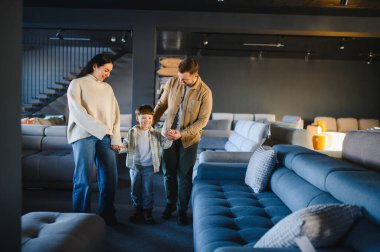 Ebeveynler ve küçük oğulları modern mobilya salonunda yürüyorlar, yeni evleri için rahat bir kanepe seçiyorlar.