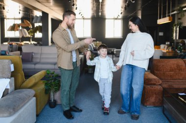 Ebeveynler ve küçük oğlu mobilya galerisinde yürüyorlar, yeni ev dekorasyonu arıyorlar, oturma odası koltukları için birlikte alışveriş yapıyorlar.