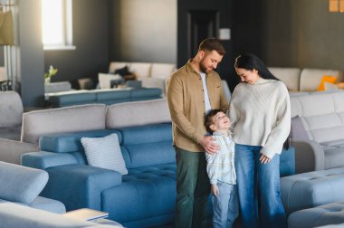 Genç bir aile, anne, baba ve oğul, yeni bir kanepe alışverişi, modern mobilya galerisinde çeşitli seçenekleri araştırıyorlar.