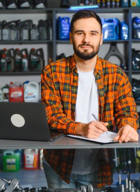 Bir oto yedek parça mağazasında çalışan sakallı bir adam, dizüstü bilgisayar kullanıyor ve bir panoya yazı yazıyor, ürünlerin raflarında duruyor.