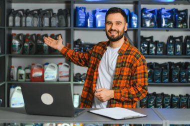 Genç, mutlu bir adam bir otomotiv dükkanında duruyor, arkasında motor yağı ve araba sıvıları sunuyor.