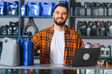Genç erkek oto yedek parça dükkânı sahibi tezgahın arkasında raflarda ve laptopta motor yağı ile gülümsüyor, envanter ve satışları yönetiyor.