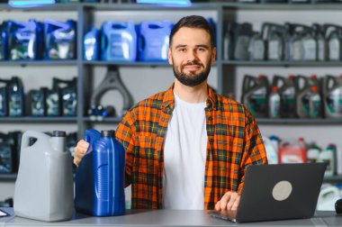 Adam gülümsüyor, oto yedek parça mağazasında motor yağı tutuyor kayganlaştırıcı ve araba bakım ürünleriyle dolu raflarda.