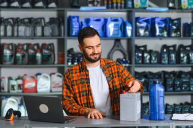 Bir oto yedek parça mağazasında çalışan gülümseyen adam, bir araba aküsü ve bir şişe motor yağı sunuyor.