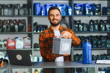 Tamirhanede çalışan sakallı arkadaş canlısı bir adam elinde araba aküsü, arka planda petrol ve otomotiv ürünleri rafları var.