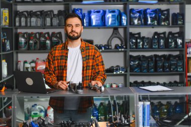 Otomobil yedek parça satan bir dükkanda kasada duran adam. Laptop, tarayıcı ve ürünlerle dolu raflar görülebilir