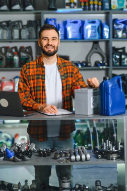 Araba yedek parça mağazası müdürü gülümsüyor, elinde araba aküsü ve motor yağı var. Arka planda çeşitli otomotiv bileşenlerine sahip raflar