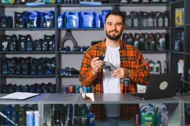 Bir otomotiv parçacıda çalışan gülümseyen tamirci, yedek parça tutuyor ve dizüstü bilgisayardaki envanteri kontrol ediyor.