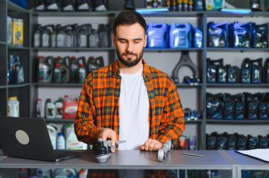 Otomotiv yedek parçalarını, arka planda kayganlaştırıcı rafları olan bir garajdaki bir masada inceleyen bir servis çalışanı.