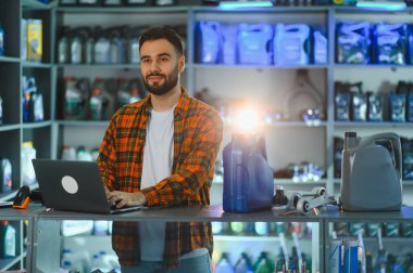 Adam oto yedek parça mağazasını yönetiyor. Tezgahta kayganlaştırıcı konteynırlar ve araba parçaları olan bir dizüstü bilgisayar kullanıyor.