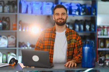 Sakallı otomobil parçaları satıcısı, karşı taramada ve satışta dizüstü bilgisayar kullanarak, küçük perakende galeride üniformalı bir şekilde gülümsüyor.