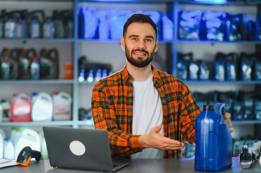 Bir oto yedek parça mağazasında çalışan gülümseyen adam, bir konteynır dolusu motor yağı sunuyor, müşterilere yardım etmeye hazır.