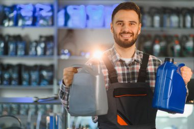 Gülümseyen tamirci motor yağı seçenekleri sunuyor, arka planda bulanık raflarla bir oto yedek parça dükkanında duruyor.