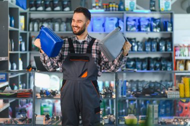 Gülümseyen tamirci bir otomotiv dükkanında farklı motor yağı şişelerini sunarak güvenilir araba bakım hizmeti sunuyor.