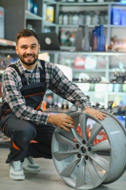 Otomobil tamircisi elinde alaşım tekerleği, garajda gülümsüyor. Profesyonel teknisyen yeni araba parçası gösteriyor