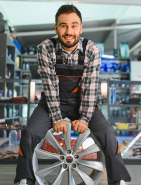 Otomobil tamircisi bir araç tamirhanesinde elinde alaşımlı tekerlekle durup, otomotiv ve bakım hizmeti sunuyor.