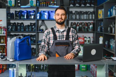Bir otomotiv yedek parça dükkanının tezgahında duran tulum giyen tamirci. Raflarda yağlar ve ürünler var.