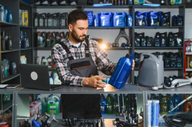 Araba dükkanı sahibi motor yağı tutuyor, garajda dizüstü bilgisayar üzerinde çalışırken araç tamiri için ürünler öneriyor.