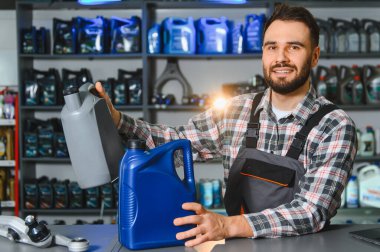 Kendine güvenen sakallı bir işçi, elinde motor yağı şişeleri, modern bir araba tamirhanesinde yedek parça ile bir rafın önünde duruyor.