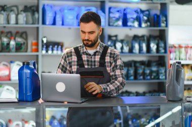 İş üniformalı teknisyen oto yedek parça mağazasında dizüstü bilgisayar kullanıyor. Kayganlaştırıcı ve araba ürünleriyle dolu raflar arka planda, envanter ve servisi gösteriyor