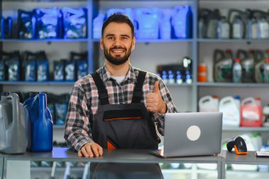 Yedek parça mağazası satıcısı tezgahın arkasında duruyor, motor yağı ve dizüstü bilgisayar sergiliyor, baş parmak işareti yapıyor.