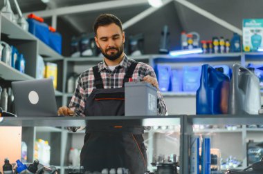 Otomotiv parçalarını yöneten erkek teknisyen, bir oto tamirhanesinde dizüstü bilgisayarla çalışırken araba aküsü tutuyor.