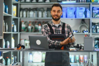 Araba parçaları dükkanında çalışırken gülümseyen bir satıcı elinde bir aletle tezgahın arkasında dizüstü bilgisayar ve araba aküsüyle dikiliyor.