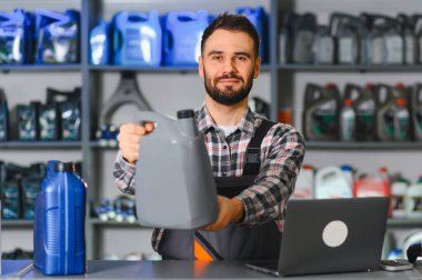 Gülümseyen tamirci bir atölyede motor yağı sunuyor, servis masasında duruyor ve çeşitli otomotiv ürünlerini raflarda sergiliyor.