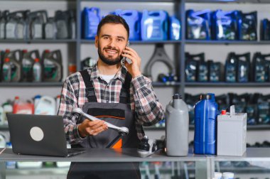 Otomobil parçaları satıcısı bir müşteriye danışıyor, elinde araba parçası, dizüstü bilgisayarı ve tezgahında kayganlaştırıcı var.