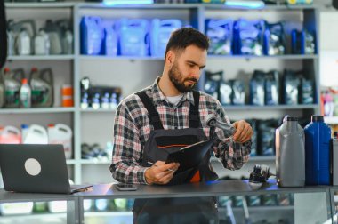 Tamir atölyesinde çalışan oto servis teknisyeni, araba parçalarını tutuyor ve bakım detaylarını içeren bir pano güncelliyor