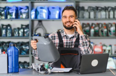 Otomobil tamircisi müşterisiyle telefonda iletişim kuruyor, elinde bir motor yağı şişesi tutuyor ve bir dizüstü bilgisayar üzerinde çalışıyor.