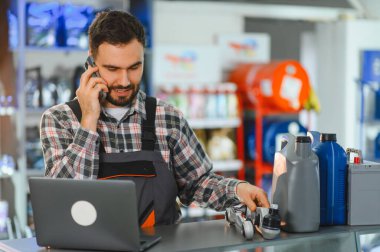 Tulum ve ekose gömlek giyen, akıllı telefonla konuşan, dizüstü bilgisayarı ve araba parçalarını kontrol eden bir erkek işçi.