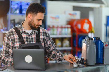 Dizüstü bilgisayar kullanan, araba parçaları, sıvı ve bileşenleri otomotiv mağazasında seçen üniformalı bir erkek tamirci.