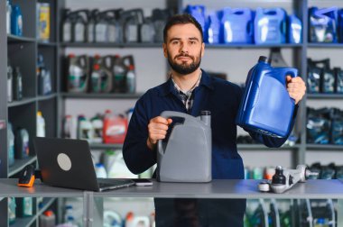 Motor yağı konteynırları tutan erkek uzman, canlı otomotiv parçaları mağazasında kameraya gülümsüyor.