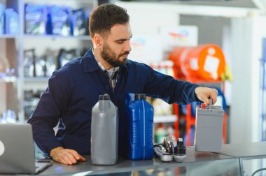 Otomotiv parçaları satan bir dükkanda araba aküsü tutan tamirci tezgahında motor yağı ve parçaları var.