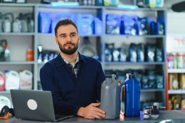Bir oto yedek parça mağazasında satış elemanı motor yağı tutuyor, tezgahta dizüstü bilgisayarın yanında duruyor.